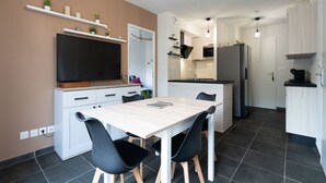 A modern kitchen with a dining area, featuring a large flat-screen TV, white cabinetry, and a tiled floor.