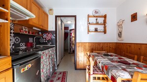 Une cuisine compacte équipée d’un réfrigérateur, d’une cuisinière et de meubles de rangement en bois. On aperçoit un coin repas avec une table et des chaises.