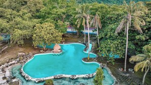 Una piscina grande y de forma irregular rodeada de rocas y vegetación, con una cabaña de techo de paja cerca.