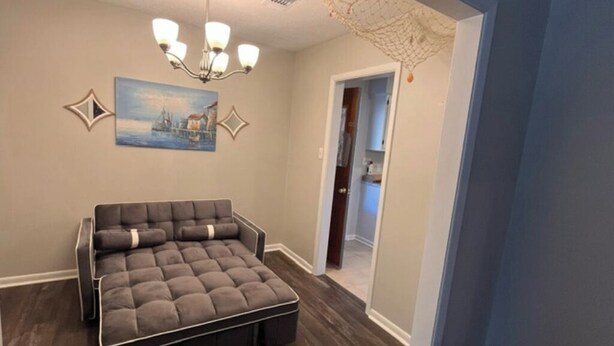A room with a large sectional sofa, a chandelier, wall art, and a doorway leading to another room. - Charming 3-bedroom house with comfortable beds close to the beach! (Long Beach)