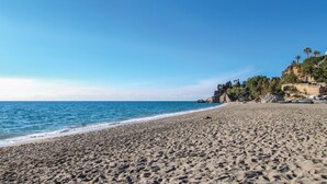 A wide sandy beach with gentle waves, a clear blue sky, and a rocky cliff with vegetation on the right. - Cozy home in Nerja with WiFi (Nerja)