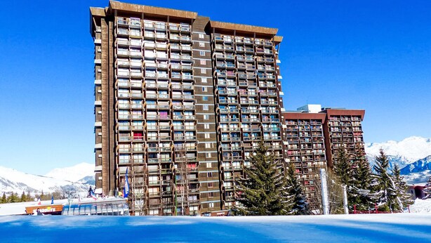 Een hotel met meerdere verdiepingen en tal van balkons, op de achtergrond een wintersportgebied en een helderblauwe lucht.