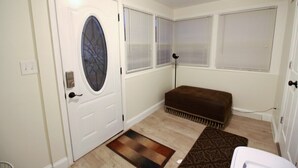 Una habitación con una puerta blanca, un tapete estampado, un banco marrón y tres ventanas con persianas.