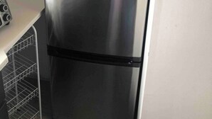 Un refrigerador de acero inoxidable con un compartimento inferior negro, situado en una cocina con gabinetes de madera y un extintor en la pared.