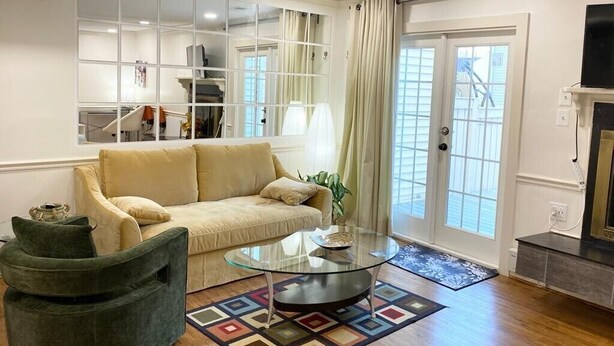 A living room with a beige sofa, a glass coffee table, and a green armchair. There is a large mirror on the wall, a television on a stand, and a fireplace. - Efficiency Bright BSMNT APT- DC 20m IAD-10m Metro (Reston)