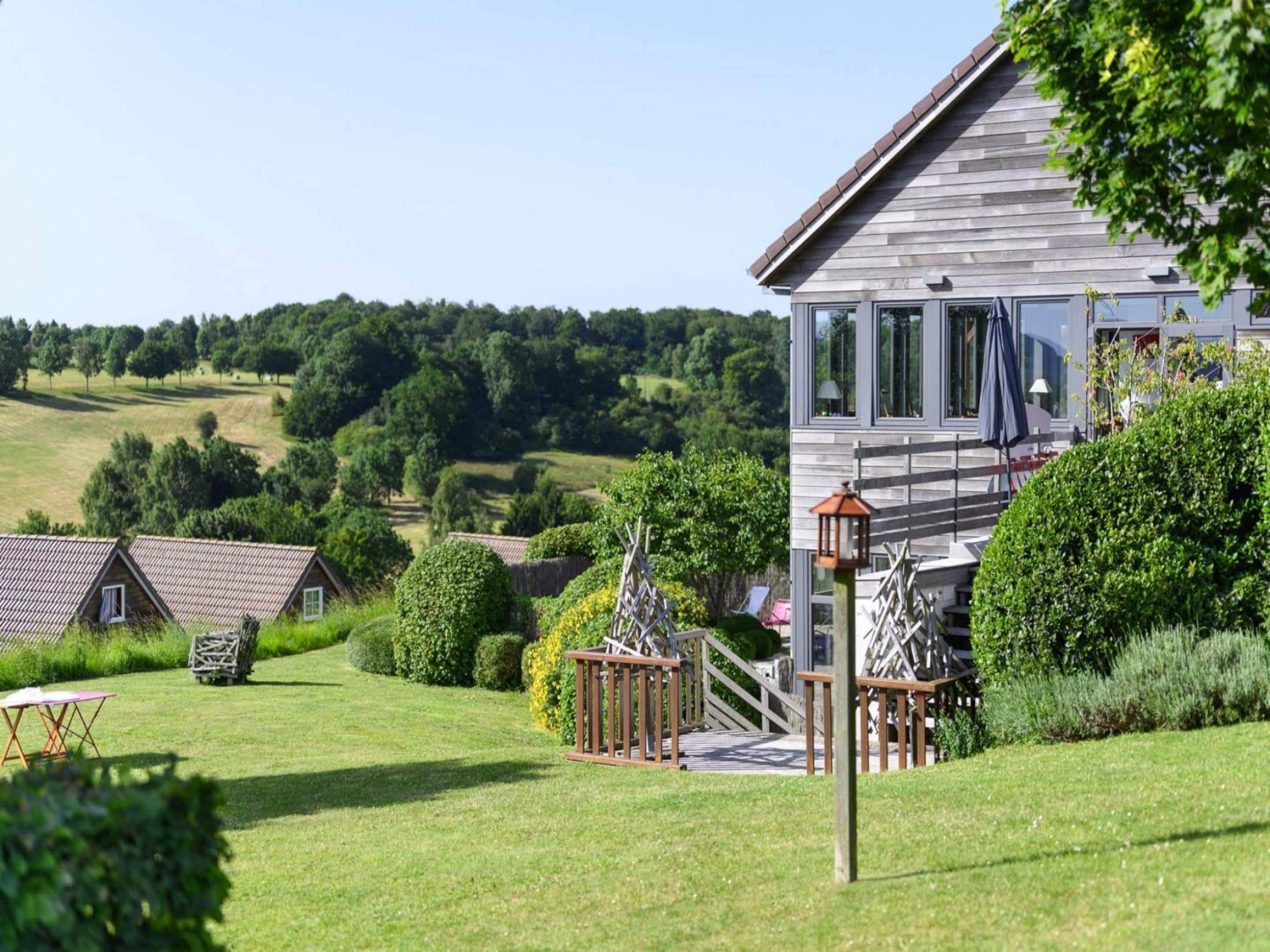 Une maison moderne dotée d’une grande terrasse, entourée de pelouses et d’arbres bien entretenus.