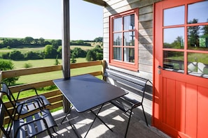 A covered outdoor seating area with a table and chairs, a red door, and a view of the countryside.