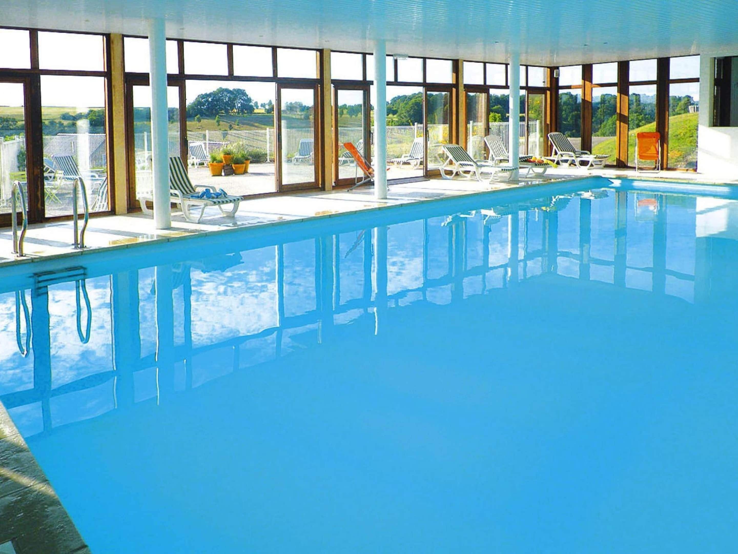An indoor swimming pool with clear glass walls, multiple lounge chairs, and a view of the surrounding landscape.