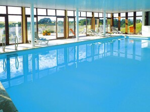 An indoor swimming pool with clear glass walls, multiple lounge chairs, and a view of the surrounding landscape. - Wooden Chalet in Abbeville Near Somme Bay (Grand-Laviers)