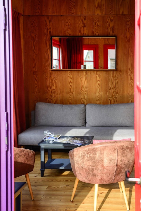 A cozy room with a wooden wall, a grey sofa, a small table with a magazine, and a pink chair.