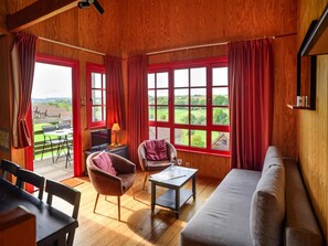Una habitación acogedora con techo de madera, ventanas grandes con cortinas rojas, un sofá, una silla, una mesita y vistas a un paisaje verde.