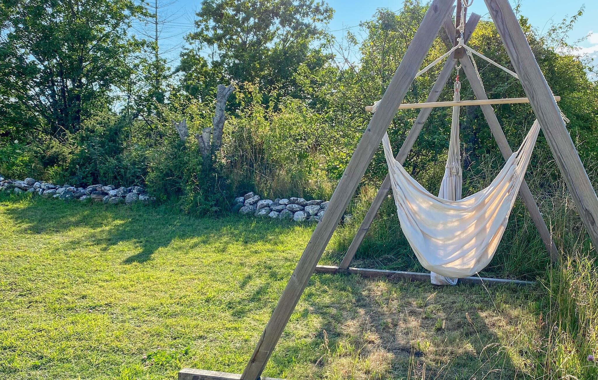 Een hangmat, opgehangen tussen houten palen, in een grasveld met bomen en een stenen muur op de achtergrond.