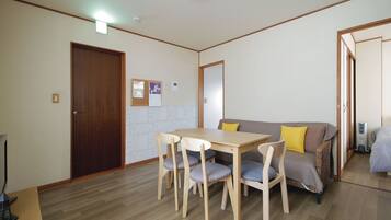 Een kamer met een houten eettafel, vier stoelen, een bank met kussens, een houten deur en een klein prikbord aan de muur.
