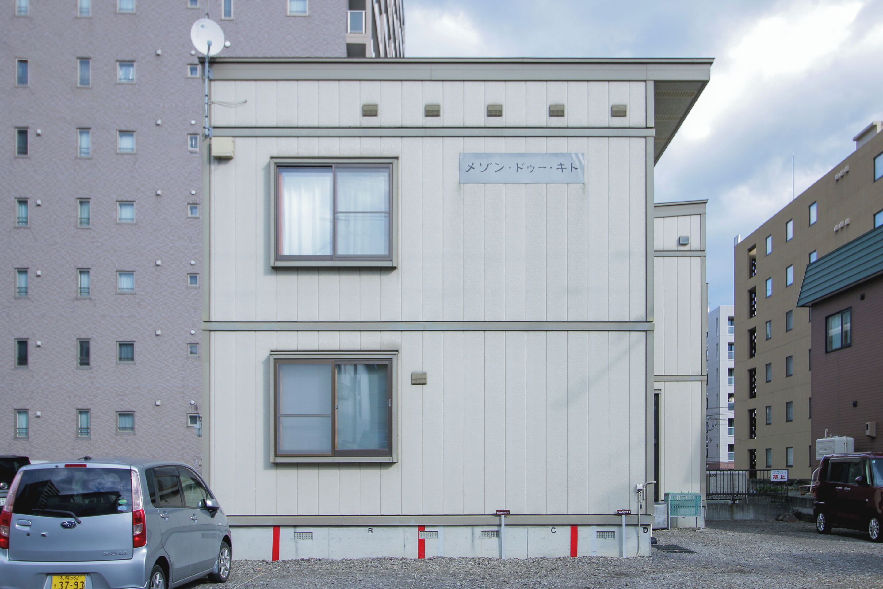 A two-story building with a sign indicating a business or office space. There are parked cars in front, and the building is adjacent to a taller residential building.