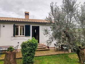 Une maison avec un toit en tuiles, une cheminée, une porte et un petit jardin avec un arbre et un arbuste.