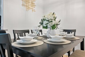 Mesa de comedor dispuesta con platos, fuentes y copas de vino blancos, rodeada de sillas negras.