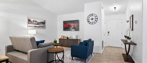 Un salon moderne avec un canapé gris, un fauteuil bleu et une table basse. Un téléviseur à écran plat affiche le logo de Netflix.