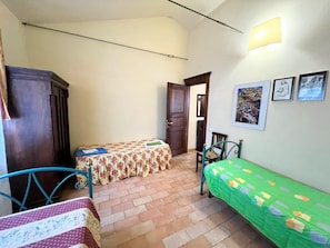 A small, narrow room with two beds, a wooden wardrobe, a door leading to another room, and a wall with framed pictures. - Apartment in Umbria Near Montefalco Winery (Giano dell'Umbria)
