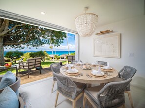 Spiseplads med et rundt bord dækket til fire, omgivet af fletstole, en lysekrone og udsigt til havet.