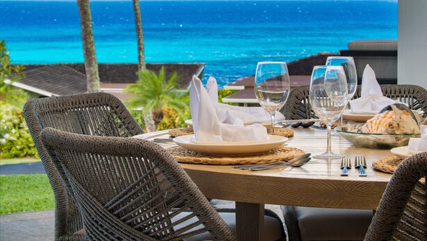 Un tavolo da pranzo apparecchiato con piatti, bicchieri e posate, con vista su un mare turchese e palme sullo sfondo.
