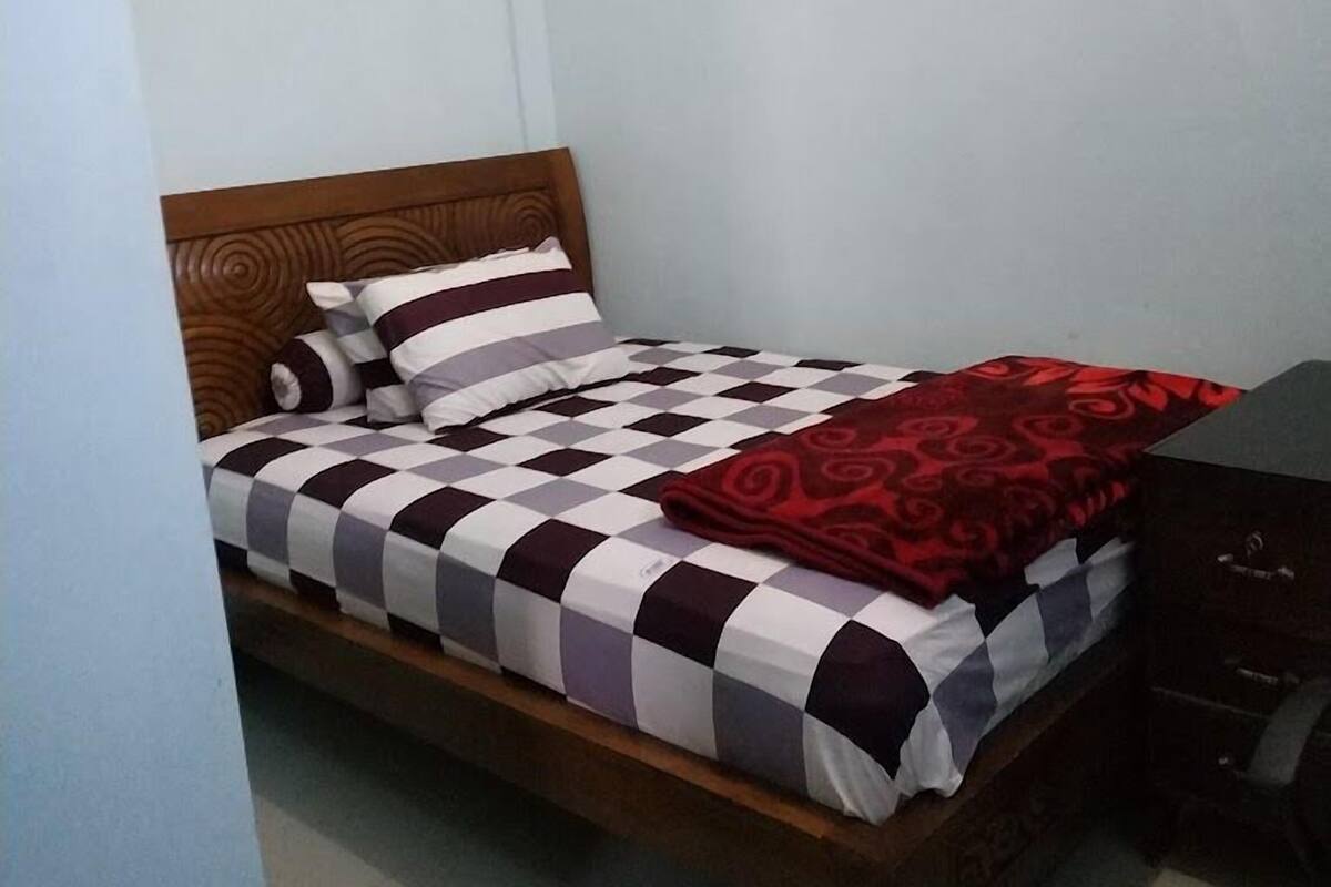 A single bed with a checkered bedspread and a red blanket, wooden headboard, and a dark-colored dresser.