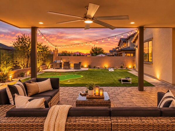 A covered outdoor patio with a ceiling fan, string lights, a lawn, and a seating area with a sofa and coffee table.