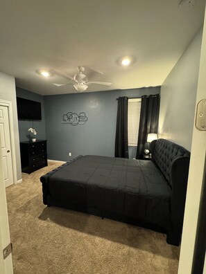 A bedroom with a bed, a ceiling fan, a wall-mounted TV, a dresser, and a window with curtains.