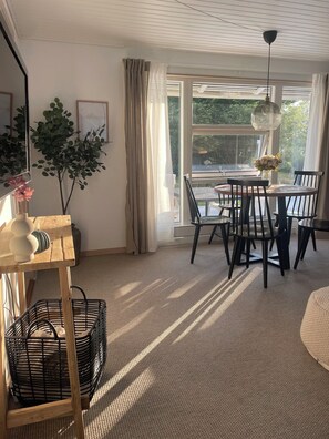 A room with a dining table, chairs, a hanging light fixture, a large window with curtains, and a plant.