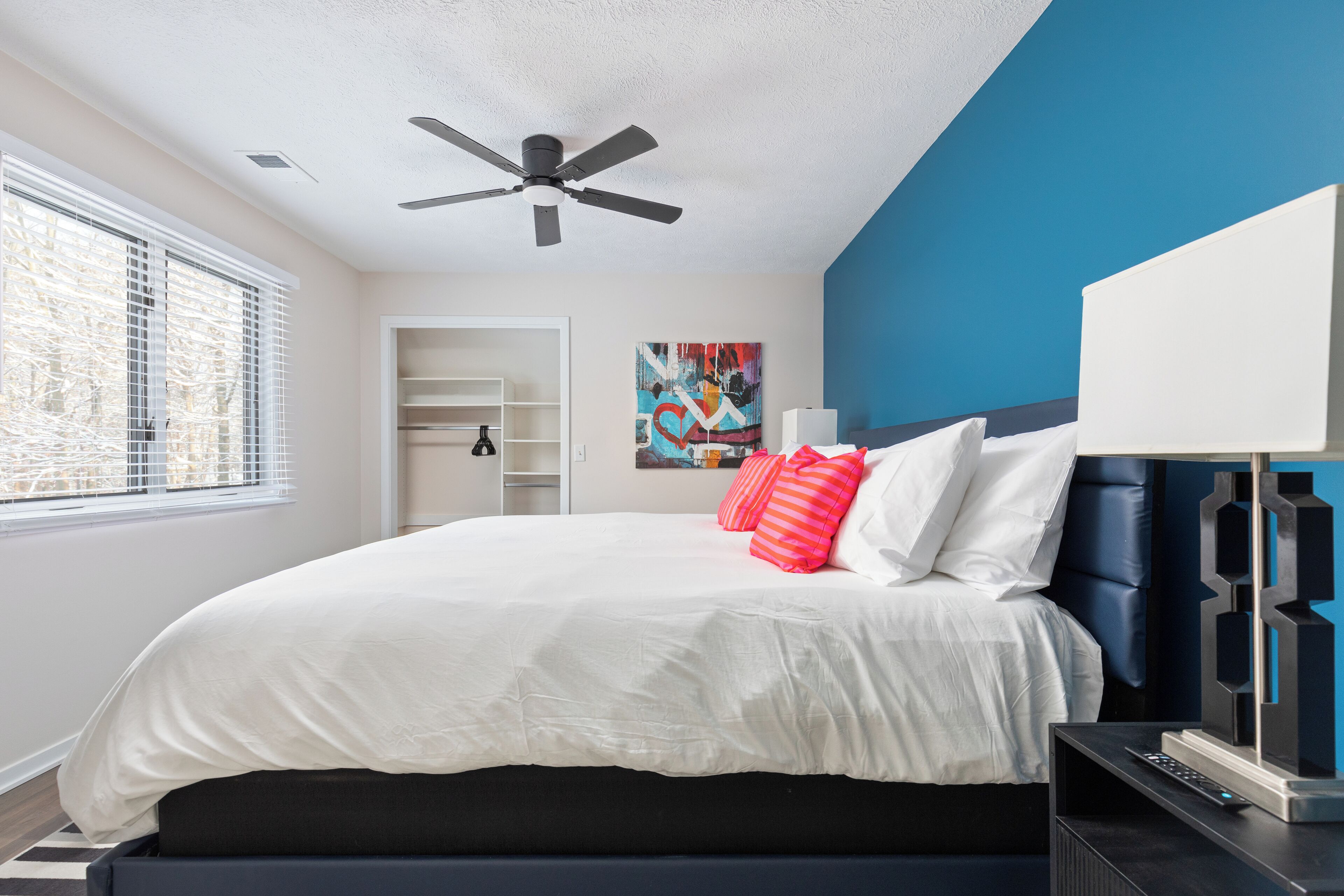 Un dormitorio con cama, ventilador de techo, mesita de noche, lámpara y un cuadro en la pared.