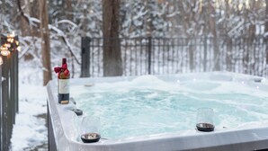 Un bain à remous avec deux verres de vin et une bouteille de vin, le tout entouré d’arbres couverts de neige.