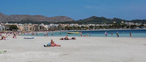 Una playa con gente tomando el sol y nadando, un bote, y edificios al fondo.