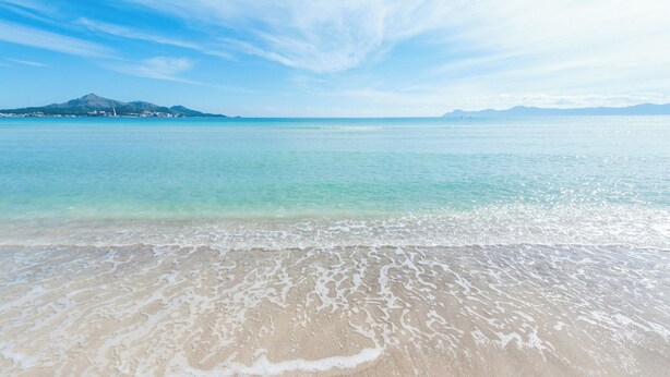 Une plage à l’eau turquoise et limpide, avec de douces vagues et une chaîne de montagnes qui se dessine à l’horizon.