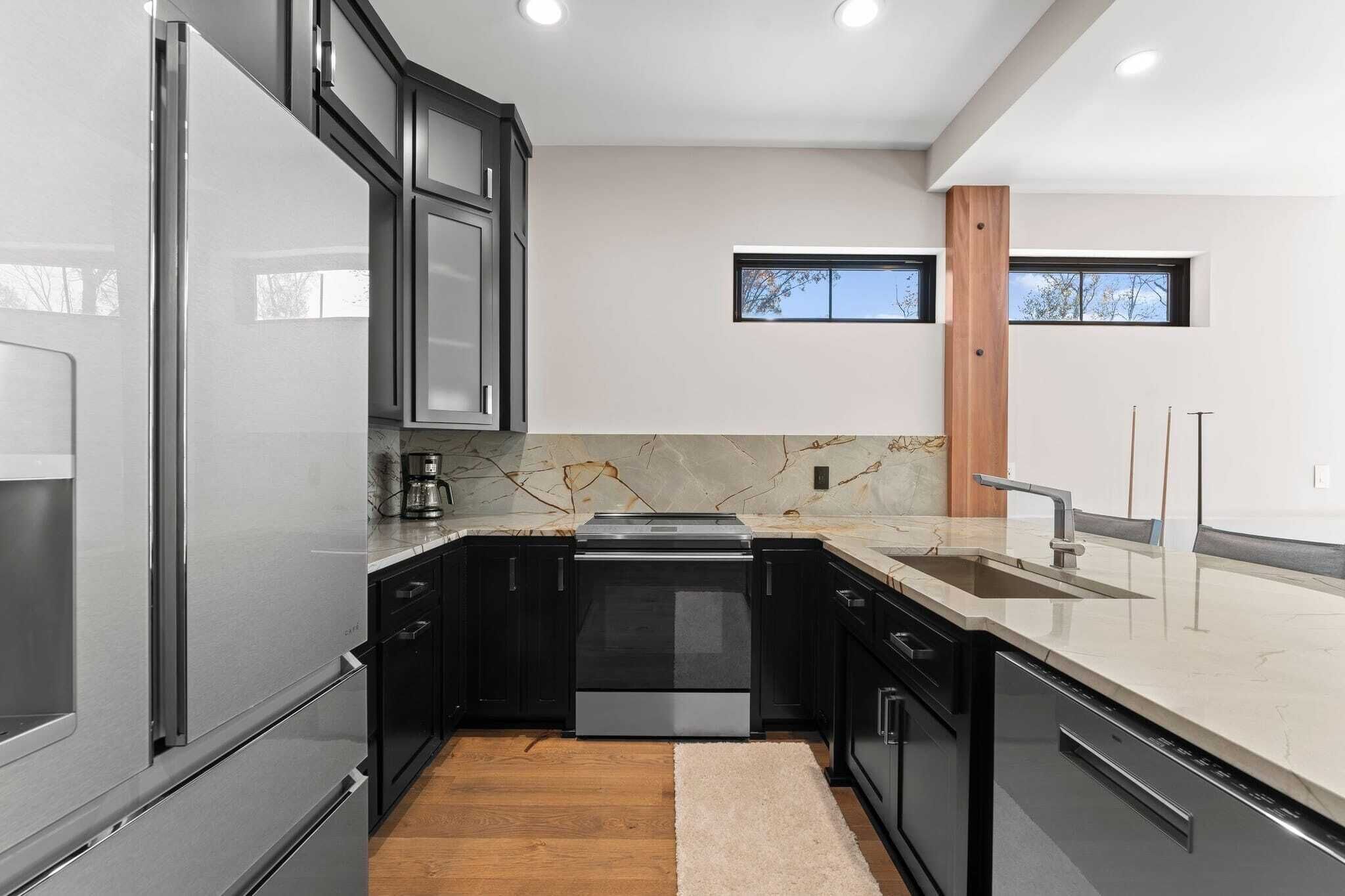 A modern kitchen with dark cabinetry, stainless steel appliances, a marble countertop, and a wooden floor.