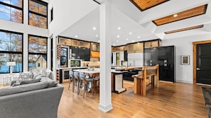 A modern kitchen with wooden cabinets, a dining area with a gray sofa, and large windows overlooking a body of water.
