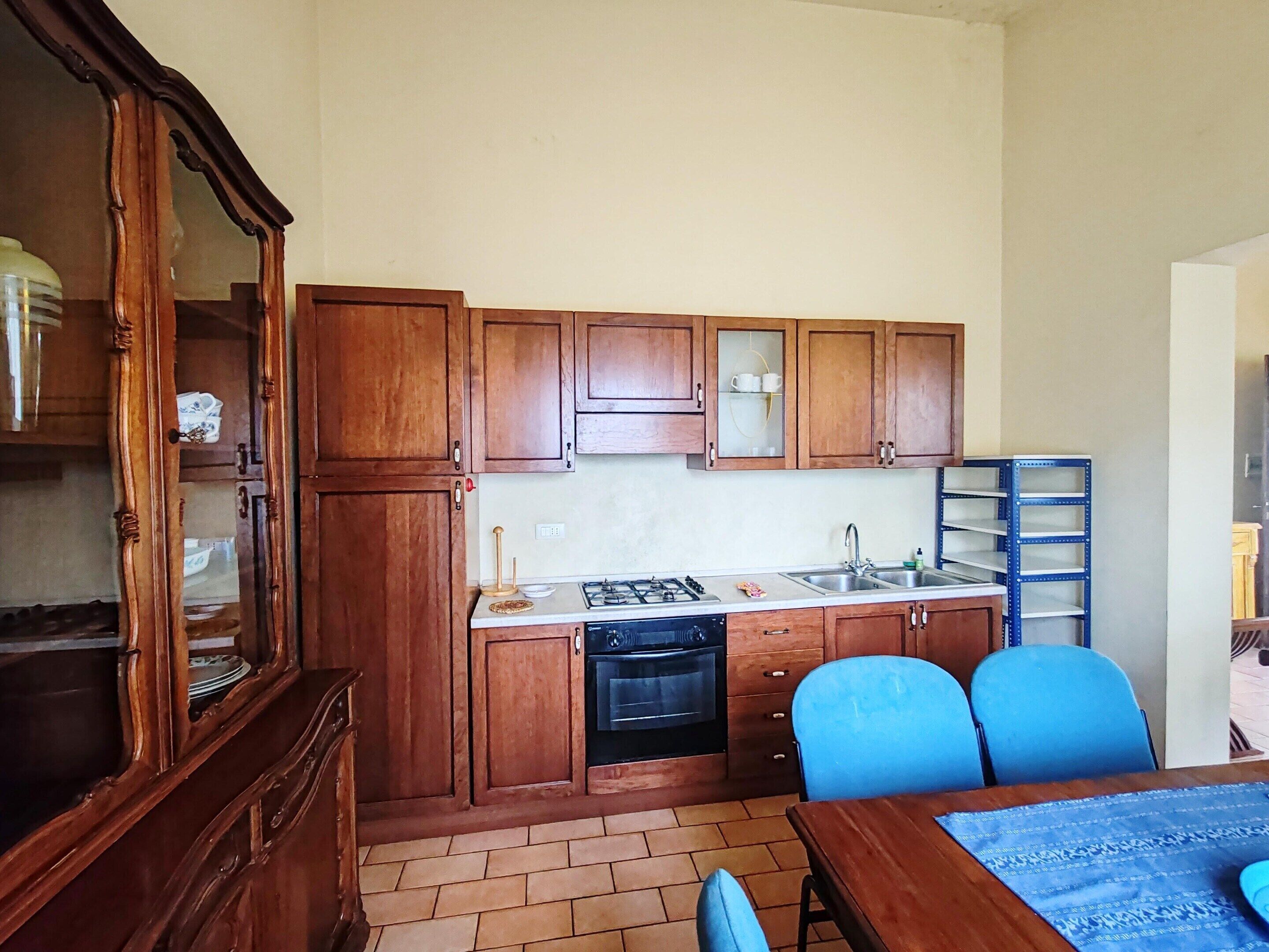 Een kleine keuken met houten kastjes, een gasfornuis, een spoelbak en een vaatwasser. Er staat een eettafel met blauwe stoelen en een blauwe tafelkleed.