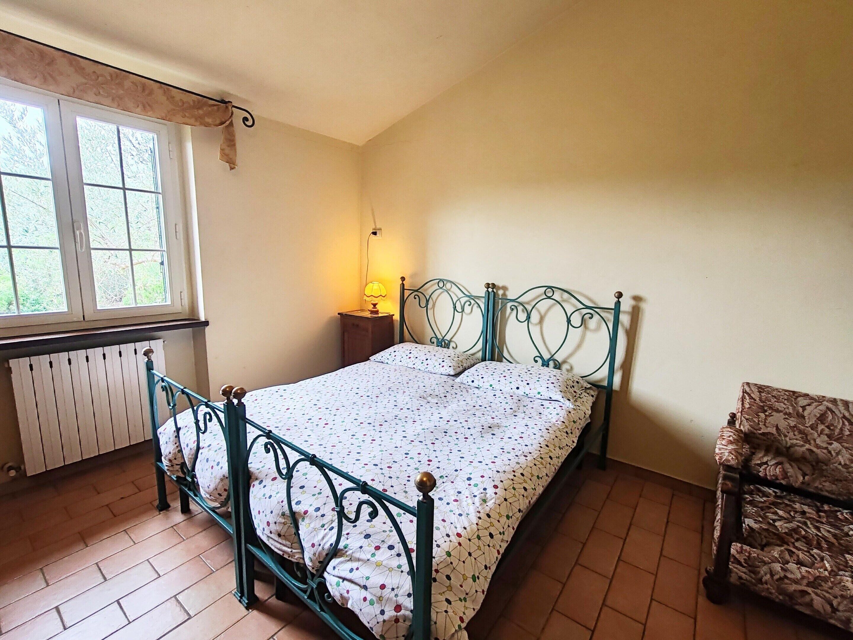 A bedroom with a bed, a nightstand, a chair, a window, and a radiator.