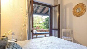 A bedroom with a bed, a chair, a small table with a lamp, and a view of a garden through an open door. - La Corte del Gallo by Interhome (Budoni)