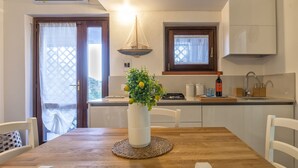 A dining area with a wooden table, white chairs, a vase with green plants, and a kitchen in the background. - La Corte del Gallo by Interhome (Budoni)