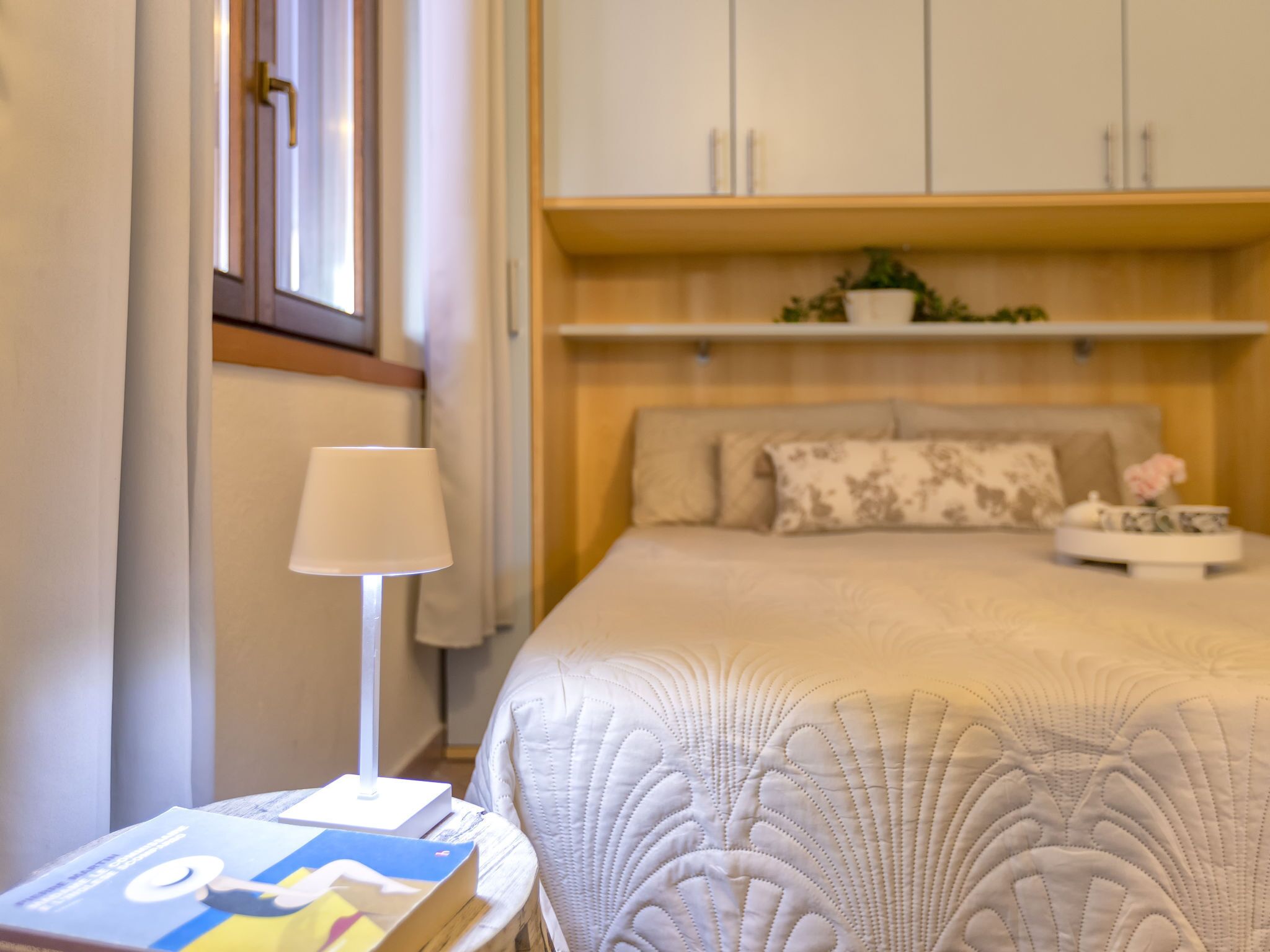 Un dormitorio ordenado con cama, mesita de noche, lámpara y un libro.