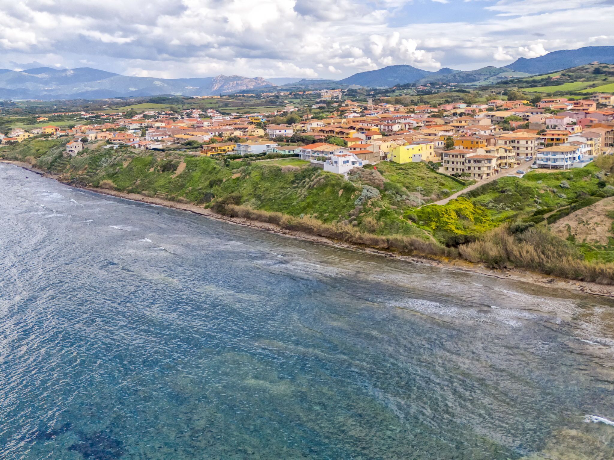 Uma vila costeira com edifícios ao longo da orla, águas azuis cristalinas e colinas verdes ao fundo.