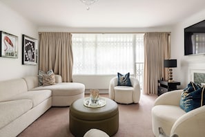 A living room with a beige sofa, a round ottoman, a chair, and a television mounted on the wall. - Extravagant 6 Bedroom Mansion by Mayfair London (London)