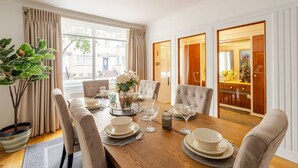 A dining room with a wooden table set for four, upholstered chairs, a chandelier, and a view of the street through large windows. - Extravagant 6 Bedroom Mansion by Mayfair London (London)