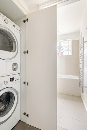 A laundry room with a front-loading washer and dryer, a white door, and a view into a bathroom with a bathtub and a window.