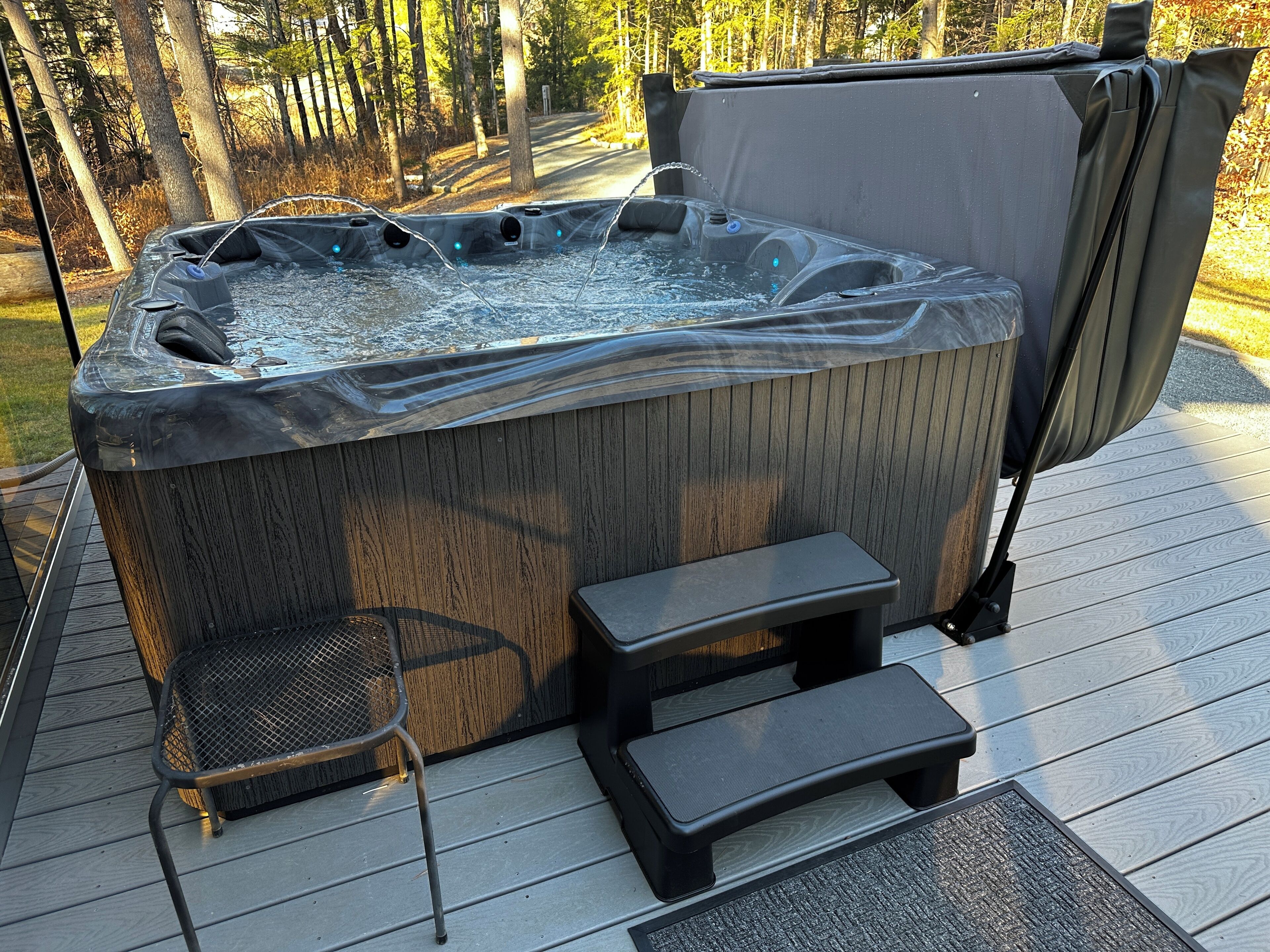 Een hottub met een houten buitenkant, een netstoel en een klein opstapkrukje op een houten terras.