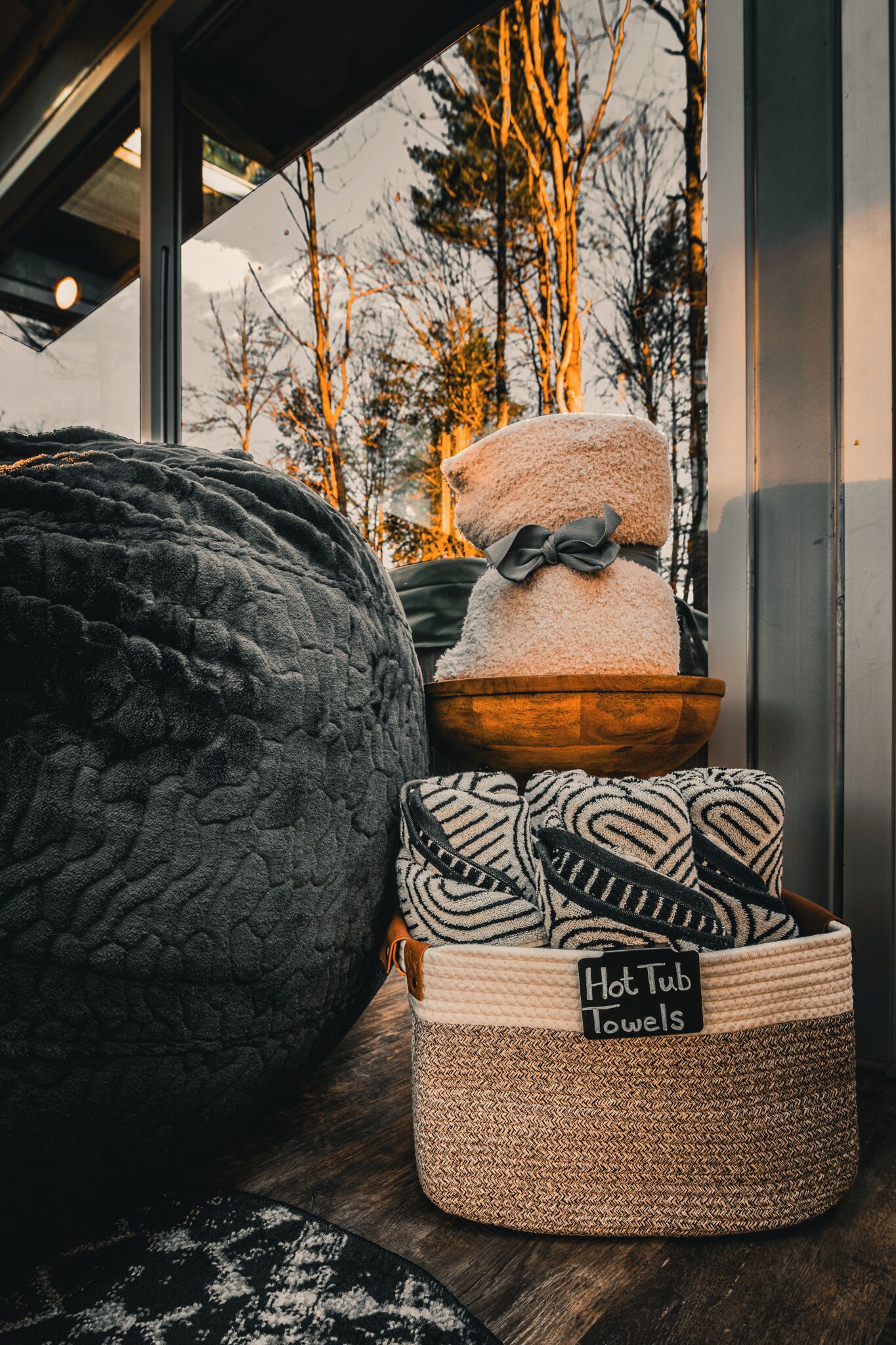 A cozy room with a large textured pillow, a woven basket labeled "Hot Tub Towels," and a bowl with a teddy bear.