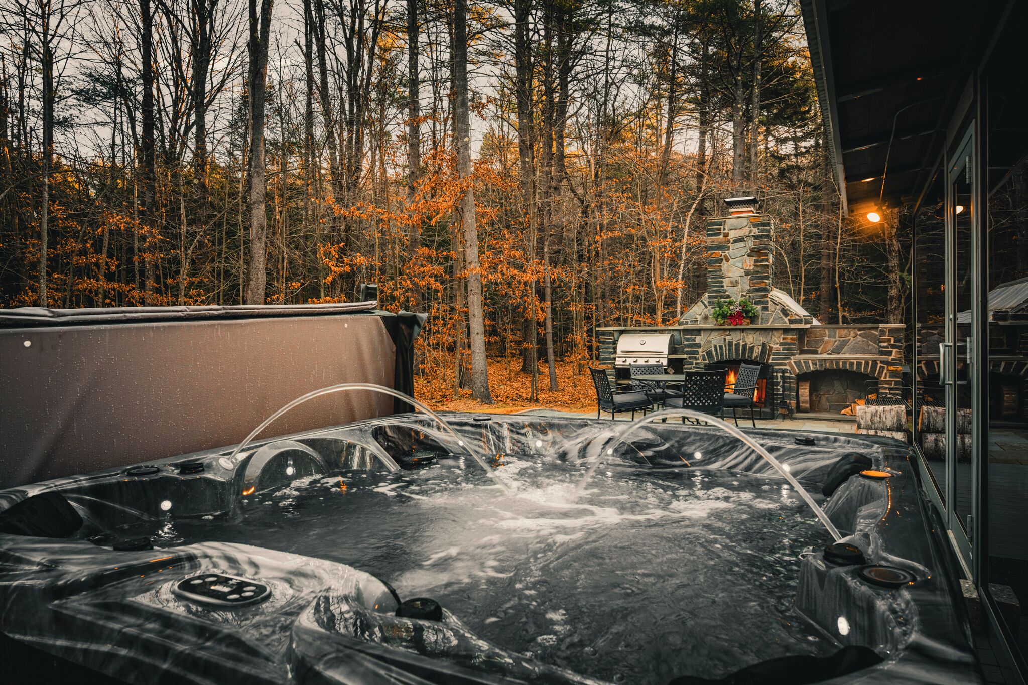 A hot tub surrounded by a forest with a stone fireplace and outdoor seating.