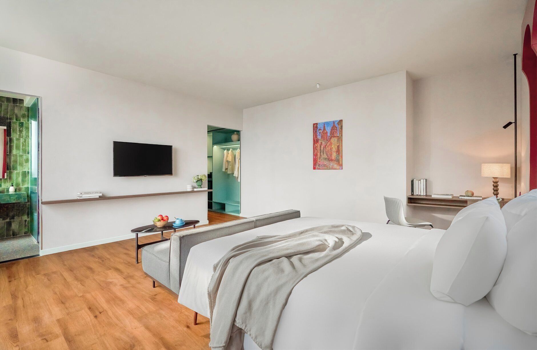 A modern hotel room with a large bed, a flat-screen TV mounted on the wall, a small table with a bowl of fruit, and a desk with a lamp.