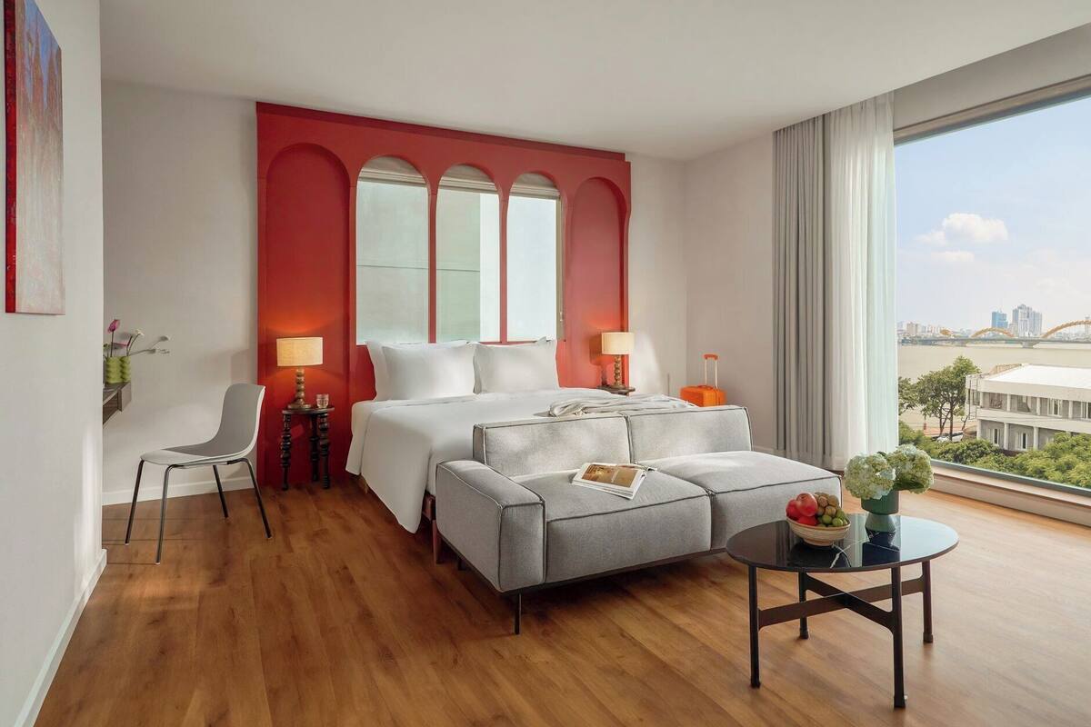Une chambre d’hôtel avec un grand lit, deux tables de chevet, un canapé gris, une petite table ronde sur laquelle se trouve un saladier rempli de fruits, et une vue sur le paysage urbain à travers de grandes fenêtres.