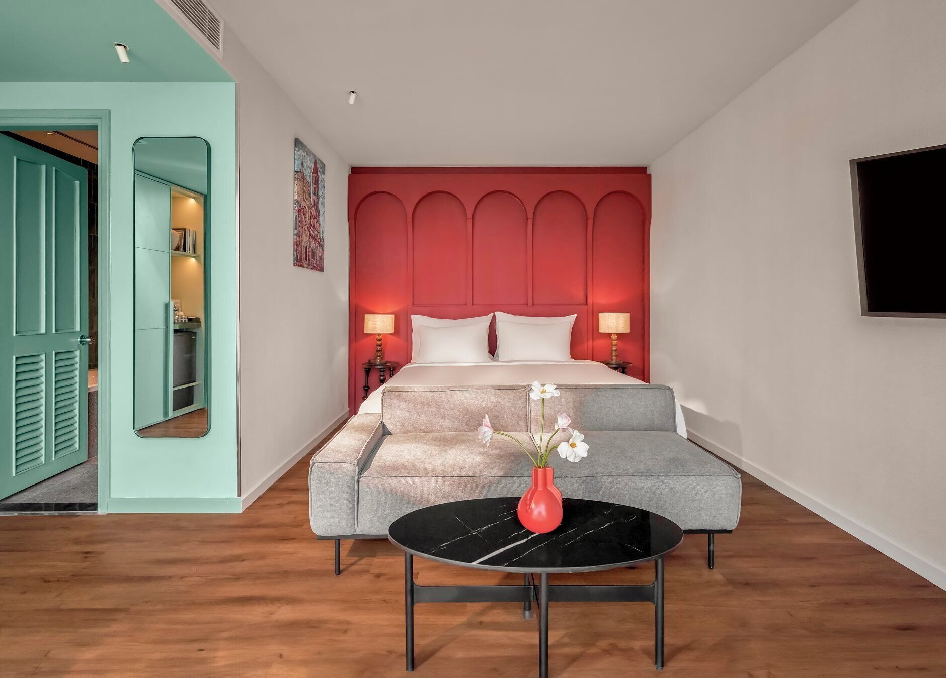 A bedroom with a bed, a grey sofa, a round table with a vase of flowers, and a red accent wall.