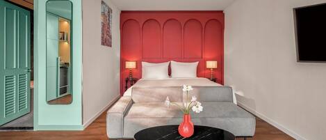 A bedroom with a bed, a grey sofa, a round table with a vase of flowers, and a red accent wall.